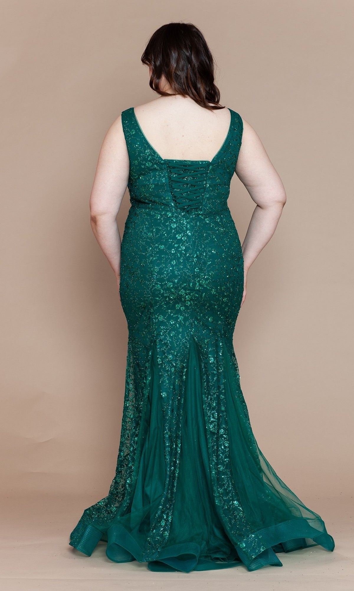 A woman stands facing away against a beige background, wearing the Plus-Size Long Glitter Mermaid Prom Dress W1136, a fitted, sleeveless emerald green gown with lace details and a flared hem.