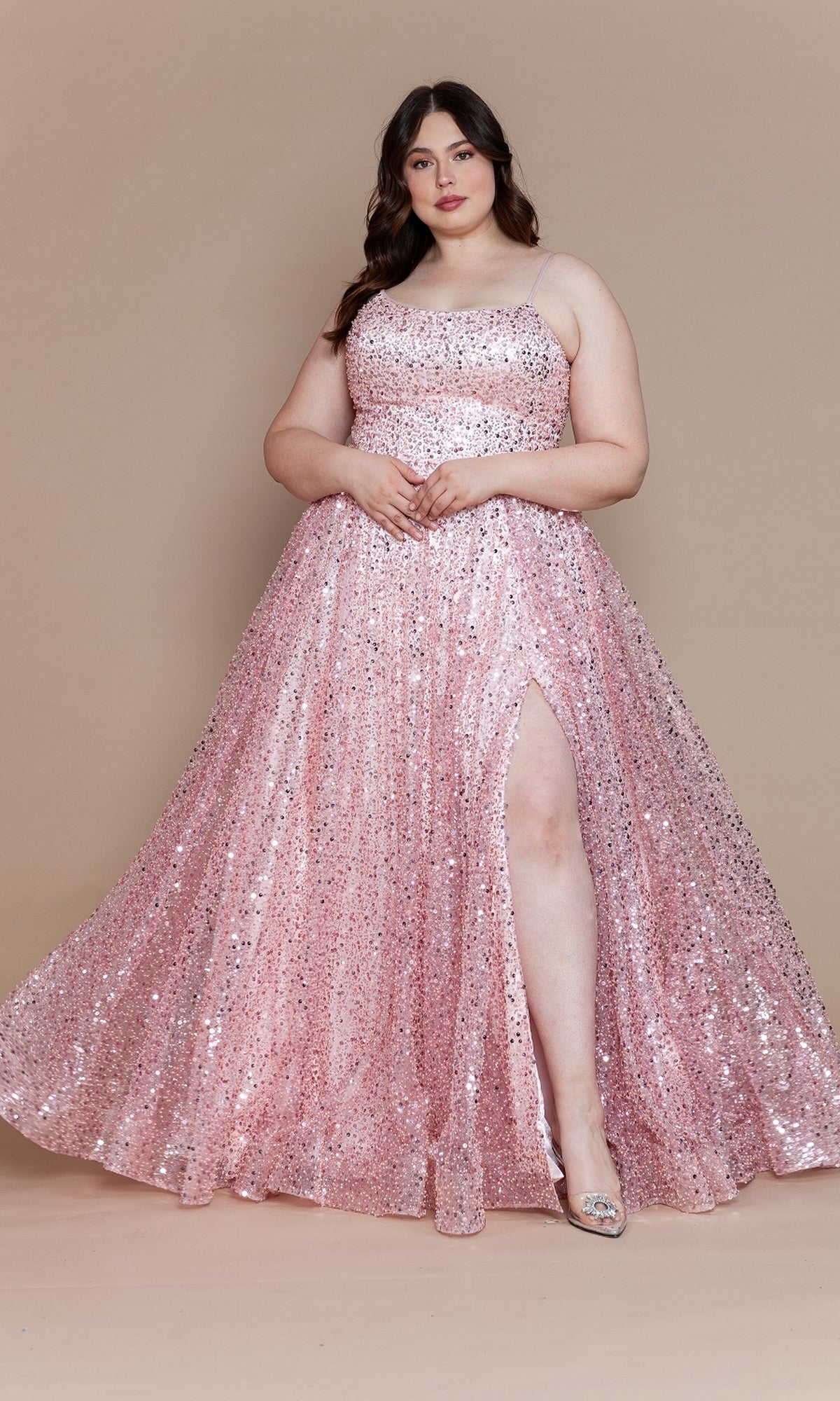 A woman poses against a beige background in the Corset-Back Plus-Size Long Sequin Prom Dress W1142 in blush, featuring spaghetti straps, a high slit, and paired with silver shoes.