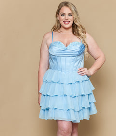 A woman wearing the Short Plus Size Homecoming Dress: Poly USA W1196—a light blue, spaghetti strap dress with a satin corset bodice and tiered ruffle details—smiles at the camera against a beige background.