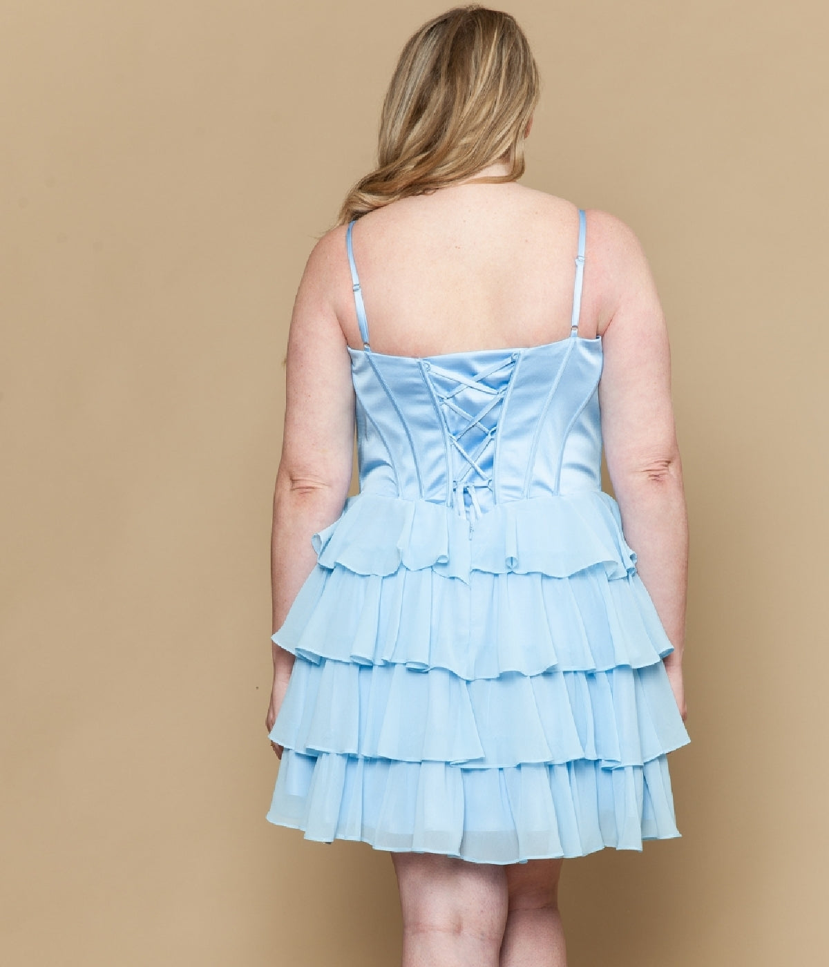 A woman in the Short Plus Size Homecoming Dress: Poly USA W1196, a light blue sleeveless dress with a ruffled chiffon A-line skirt and lace-up back, stands facing away against a beige background.