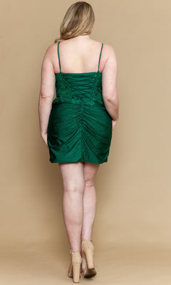 A woman in the green Poly USA W1200 short plus size homecoming dress with a ruched corset bodice and beige heels stands with her back to the camera against a beige background.