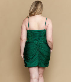 A person with light skin and blonde hair faces away, modeling the Poly USA W1200 Short Plus Size Homecoming Dress—a green sleeveless dress with lace appliques, ruching, and a delicate corset bodice—against a beige background.