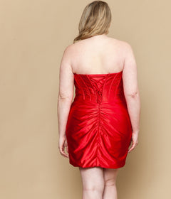 A woman models the Poly USA w1204, a plus-size short semi-formal dress in fitted red with ruched detailing and lace-up back, standing facing away against a plain beige background.