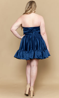 A woman models the Poly USA W1212 Short Plus Size Homecoming Dress, a navy blue strapless satin gown with a ruffled hem and fit-and-flare silhouette, standing against a beige background.