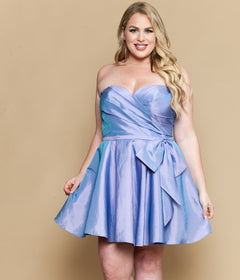 A woman smiles at the camera in the Poly USA W1214, an affordable plus-size strapless cocktail dress in lavender, featuring a fit-and-flare silhouette and bow detail, standing against a beige background.
