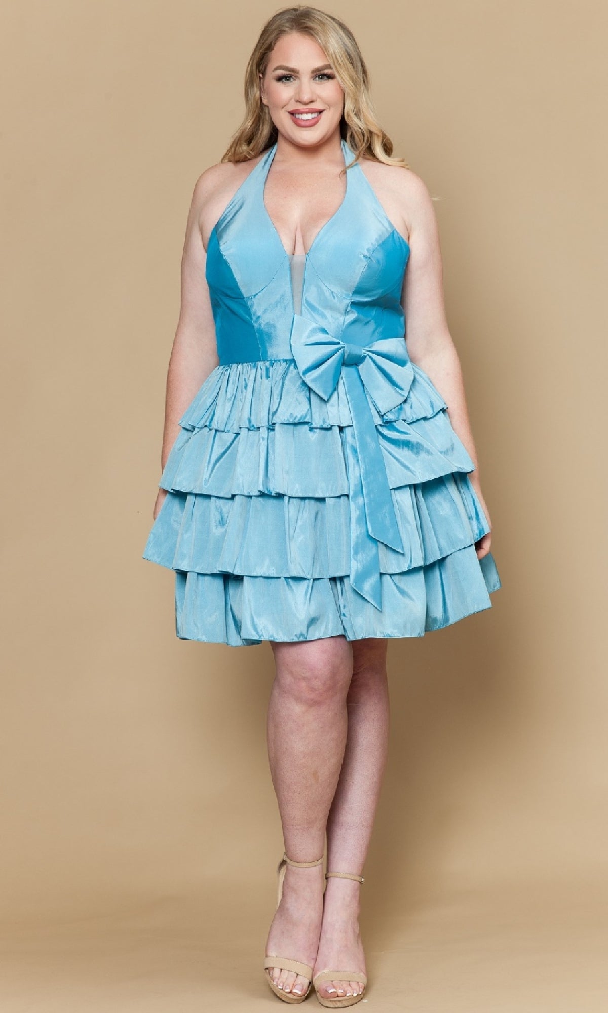 A woman poses against a beige backdrop wearing the Poly USA W1216 plus-size short halter party dress in light blue, featuring layered ruffles and a large bow, styled with nude heels.