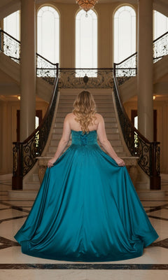 A woman in the Long Plus-Size Prom Dress: Poly USA W1244, a shimmering strapless teal satin gown, stands at the base of an ornate staircase in a grand foyer, facing away from the camera.
