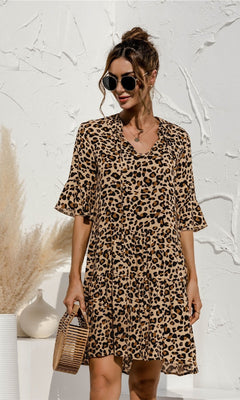 A woman wearing the Tiered A-Line Short Sleeve Casual Print Dress and sunglasses holds a small bamboo handbag, standing by a textured white wall with pampas grass in a vase nearby.