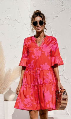 A woman wearing the Tiered A-Line Short Sleeve Casual Print Dress, sunglasses, and holding a wooden handbag stands before a white textured wall with pampas grass in a white vase beside her.