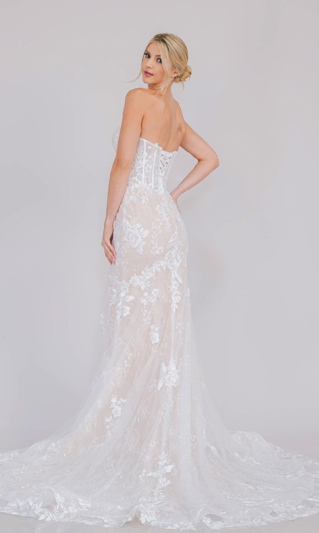 A woman stands sideways, wearing the Long White Bridal Gown: Amelia Couture WL0100, featuring delicate floral patterns, white lace, and a long train against a plain light background.