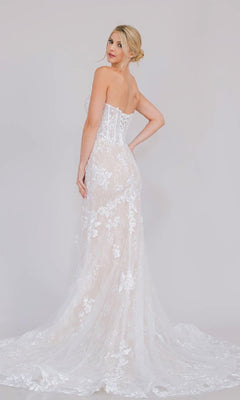 A woman stands sideways, wearing the Long White Bridal Gown: Amelia Couture WL0100, featuring delicate floral patterns, white lace, and a long train against a plain light background.