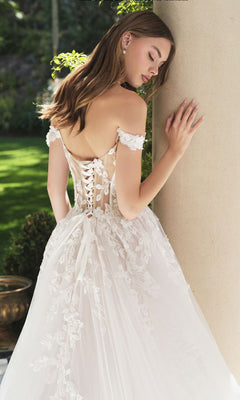 A woman in the Andrea and Leo WL057 long wedding dress, featuring an off-shoulder design with a lace-up back and floral embroidery, stands by a column outdoors, highlighting the elegance of this stunning gown.