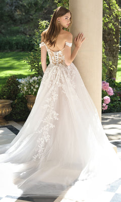 A woman wears the Andrea and Leo WL057 long wedding dress with a lace-up back and floral details, standing outdoors near a column and garden, facing away.