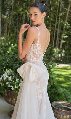A woman in the Long Wedding Dress: Andrea and Leo WL068 with a lace design, sheer corset bodice, low back, and large bow stands outdoors near greenery, looking over her shoulder.