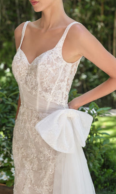 A woman in the Andrea and Leo WL068 long wedding dress—white lace with a sheer corset bodice, beaded straps, and a large tulle bow at the hip—stands outdoors amid lush greenery.