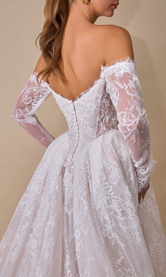 A woman wearing the Andrea & Leo WL079 long wedding dress, an elegant off-the-shoulder bridal gown with long lace sleeves and a buttoned back, stands against a plain beige background.