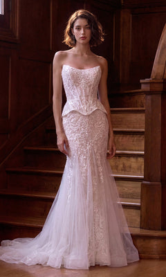 A woman wearing the Andrea & Leo WL091 long wedding dress in off white—featuring a strapless, fitted lace silhouette with a sheer bodice and elegant mermaid skirt—stands in front of a wooden staircase, gazing at the camera.