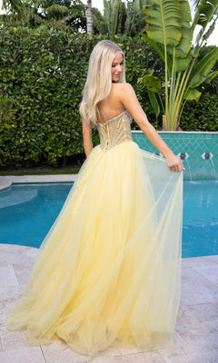 A woman with long blonde hair stands poolside wearing the Portia and Scarlett XO047, a strapless tulle ball gown with a beaded corset bodice and sheer back. She holds the flowing skirt as green foliage frames this elegant scene.