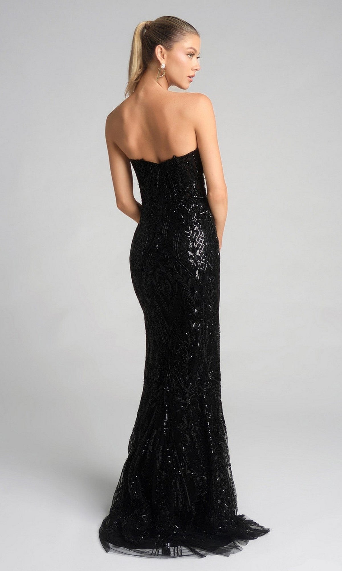 A woman faces away, wearing the Portia and Scarlett XO134 long formal dress with a fitted silhouette and sparkling embellishments, her hair in a ponytail, standing against a plain light background.