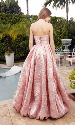 A woman in the Portia and Scarlett XO180 long formal dress, featuring a strapless a-line silhouette with a boned corset bodice and pink textured fabric, stands poolside with her back to the camera, surrounded by greenery and outdoor furniture.
