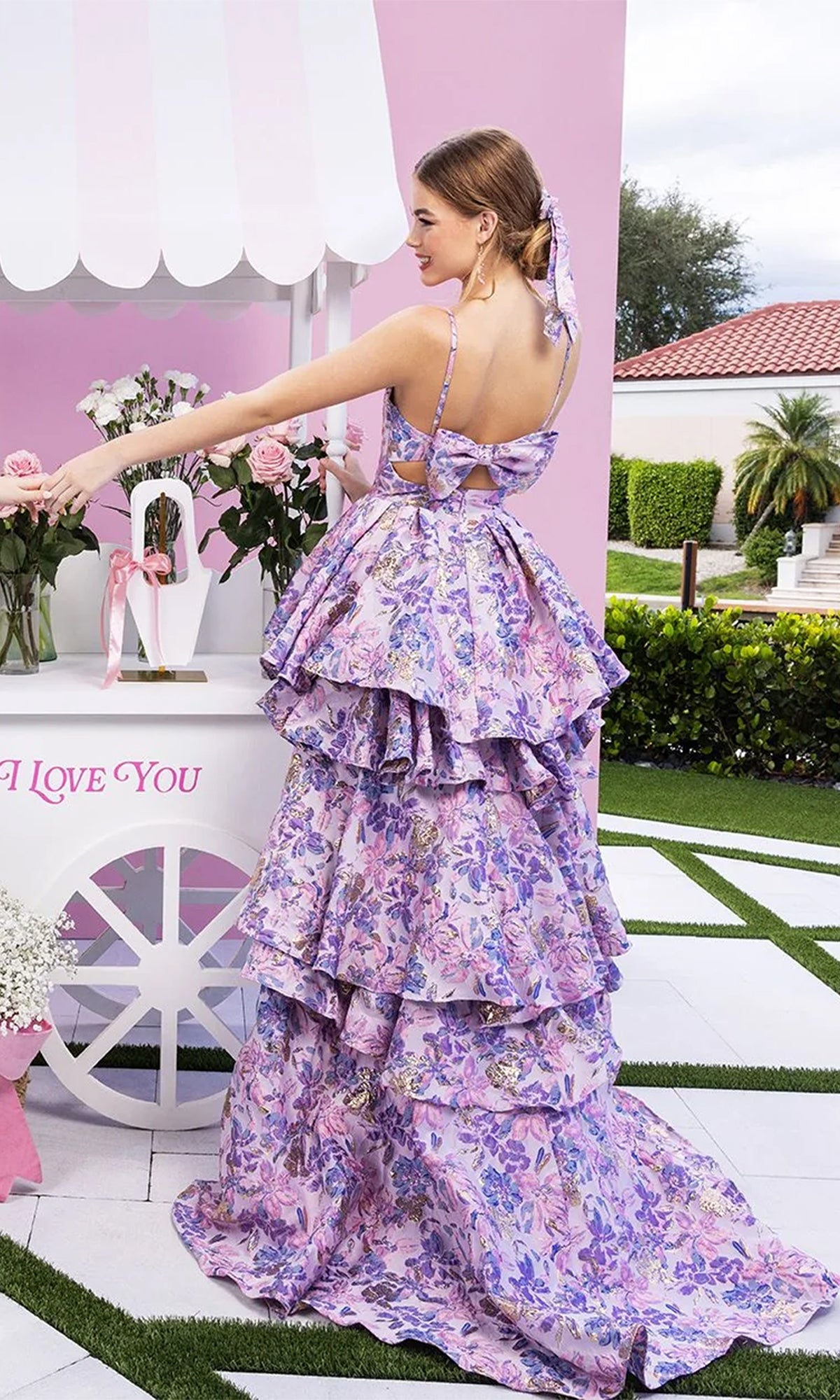 A woman in a lilac Portia and Scarlett XO181 long formal dress stands outdoors next to a white cart adorned with pink roses and a sign that reads 
