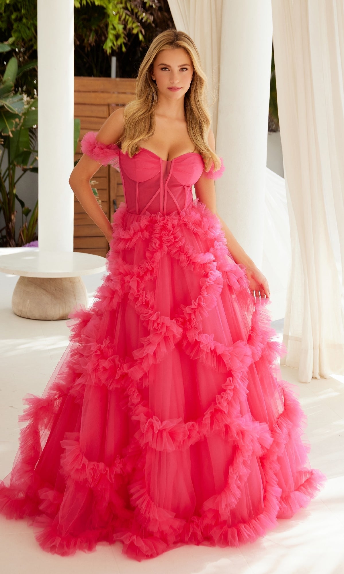 A woman stands indoors in the Sheer-Waist Off-Shoulder Long Prom Ball Gown Y1472, a bright pink dress with ruffled tulle. White columns, curtains, and greenery are visible in the background.