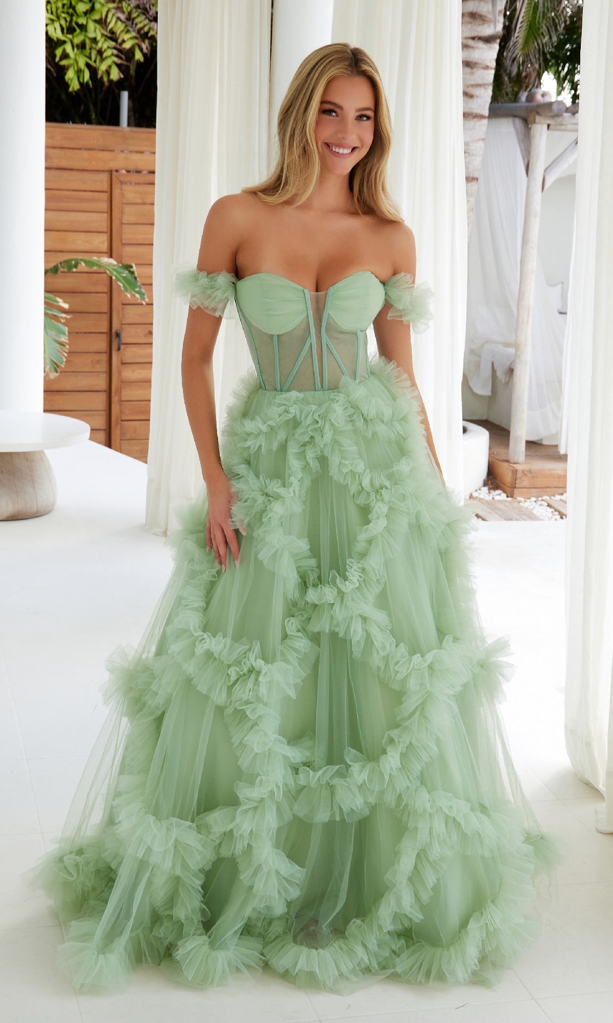 Indoors, a woman wears the Sheer-Waist Off-Shoulder Long Prom Ball Gown Y1472 in light green, featuring ruffled tulle, a fitted semi-sheer bodice, and a full skirt.