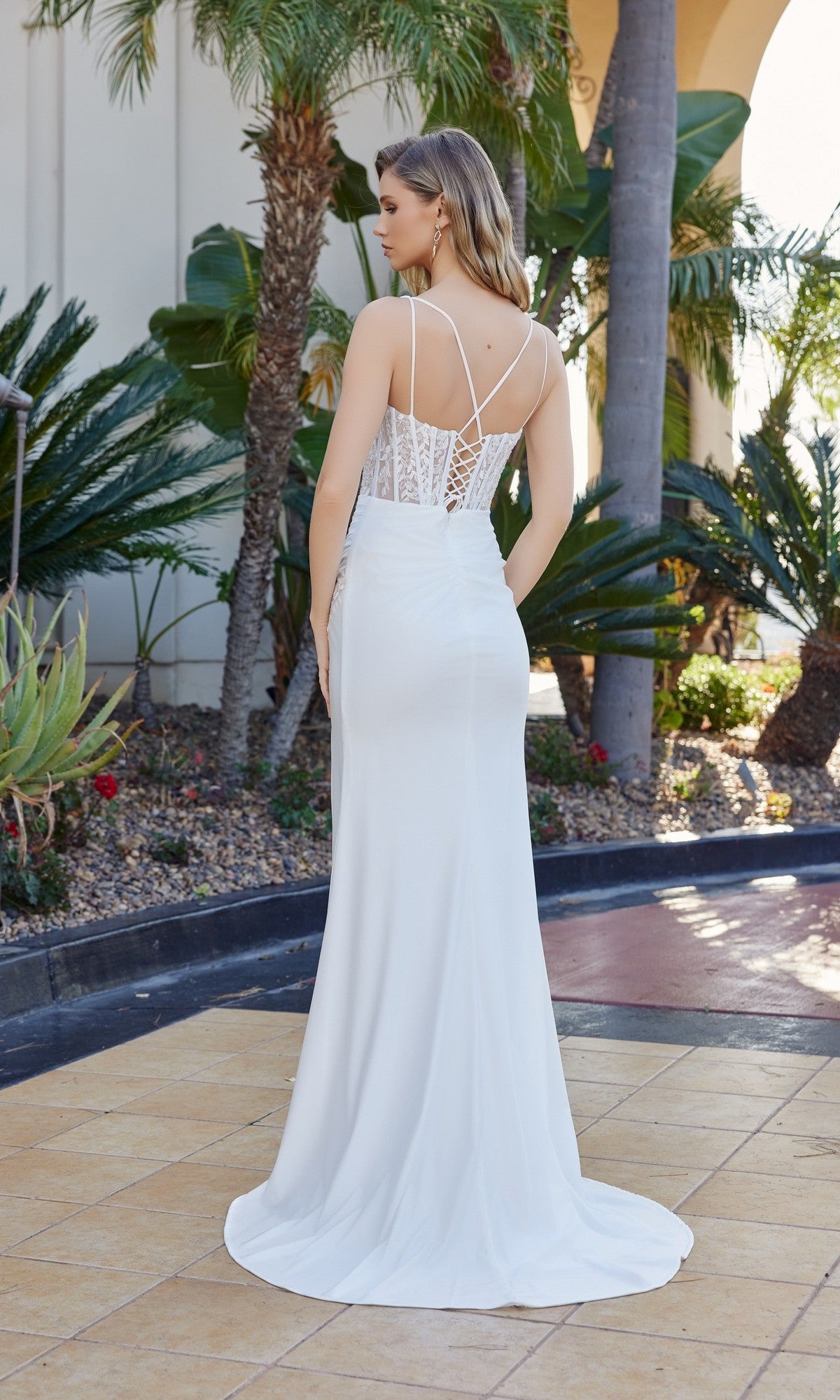 A woman stands outdoors among tropical plants, wearing the Nox Anabel Y1550 Lace-Corset Long Prom Dress, a white floor-length gown with a lace back and thin straps.