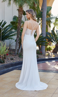 A woman stands outdoors among tropical plants, wearing the Nox Anabel Y1550 Lace-Corset Long Prom Dress, a white floor-length gown with a lace back and thin straps.