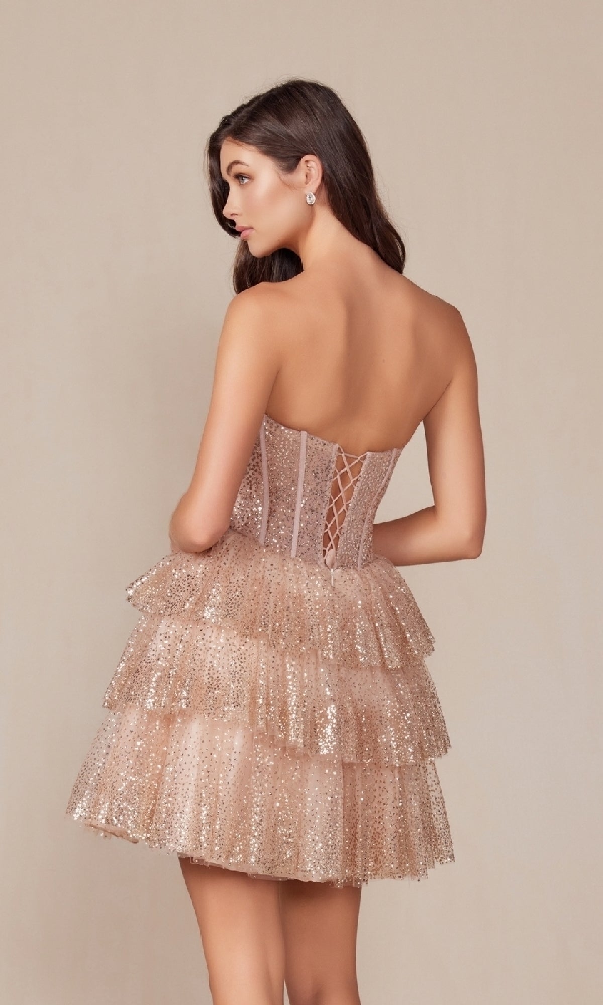 A woman stands facing away from the camera against a plain background, modeling the Strapless Short Champagne Glitter Hoco Dress Y863 with a corset-style bodice and tiered glittery skirt.