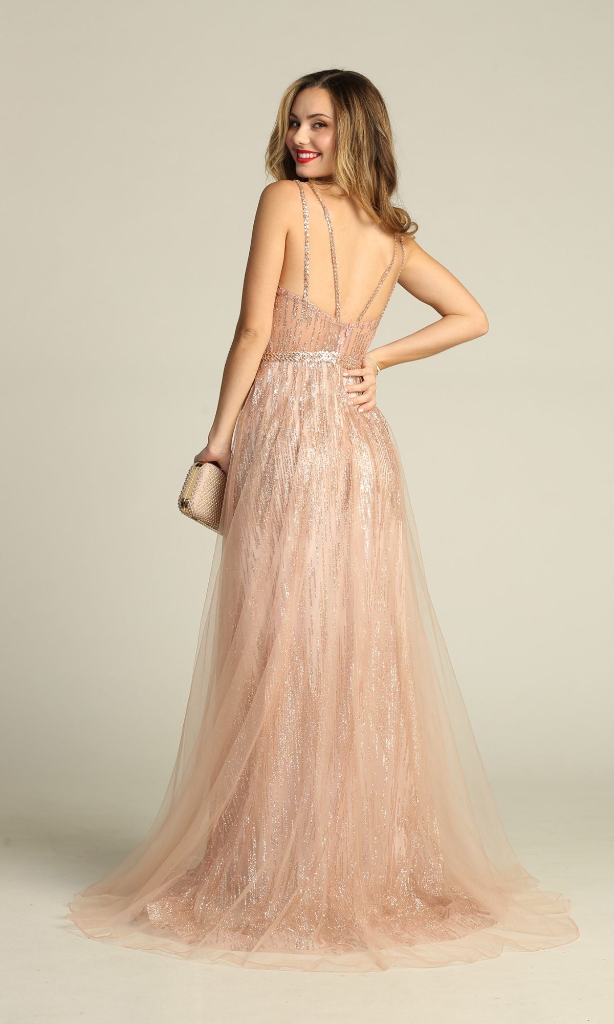 Woman facing backward in the Sheer-Waist Long Sequin Prom Dress (Chicas YG5021), a sleeveless blush gown with tulle overlay, holding a clutch and smiling over her shoulder.