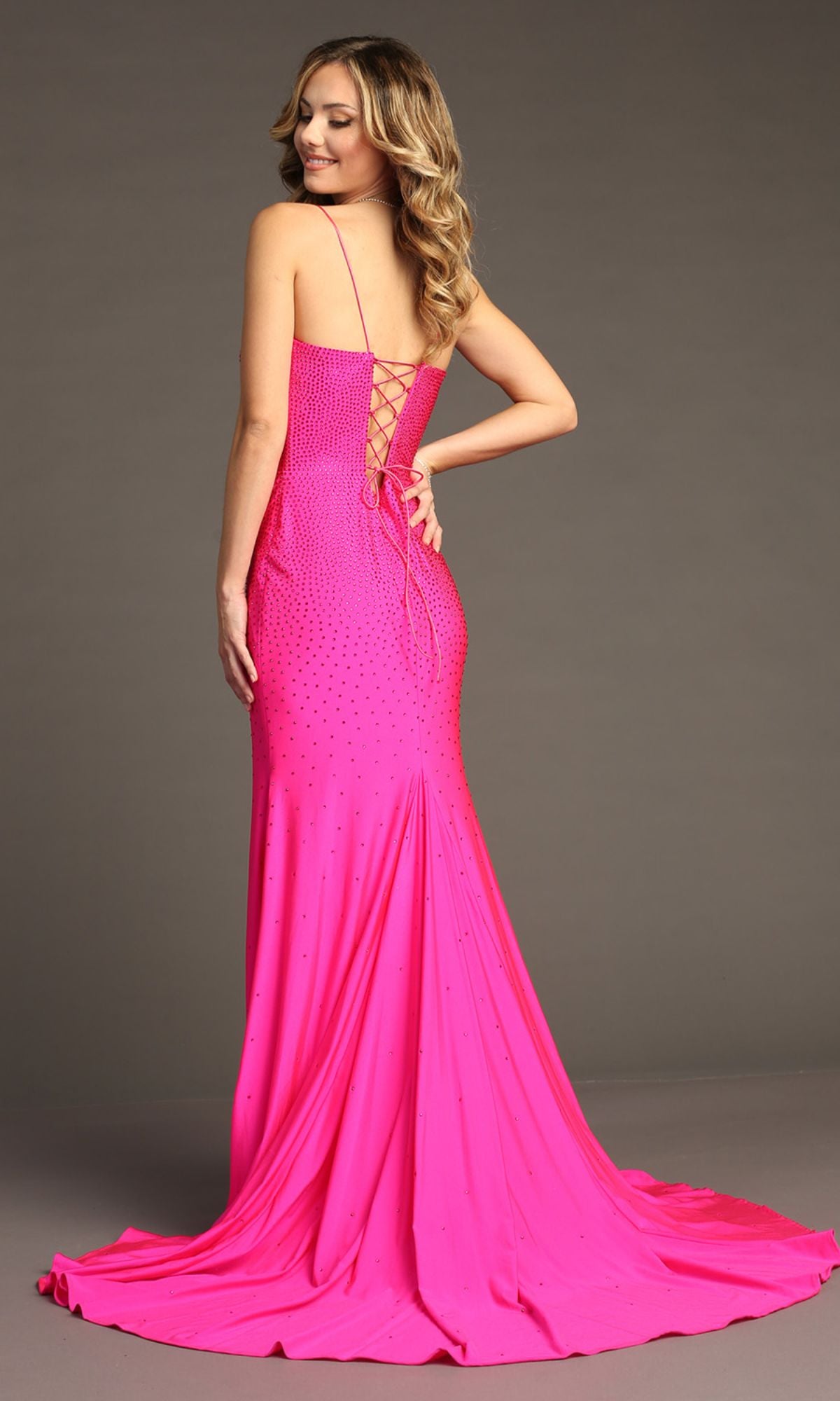 A woman models the Chicas YG5026 Lace-Up Long Beaded Prom Dress, featuring a bright pink hue, fitted bodice, lace-up back, and delicate embellishments, standing against a plain gray background.