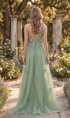 A woman with wavy blonde hair stands outdoors in a garden, wearing the Long Prom Dress: Chicas YG5028—a light green, one-shoulder gown adorned with delicate floral appliqués and featuring a lace-up back, complemented by an elegant A-line glitter tulle skirt.