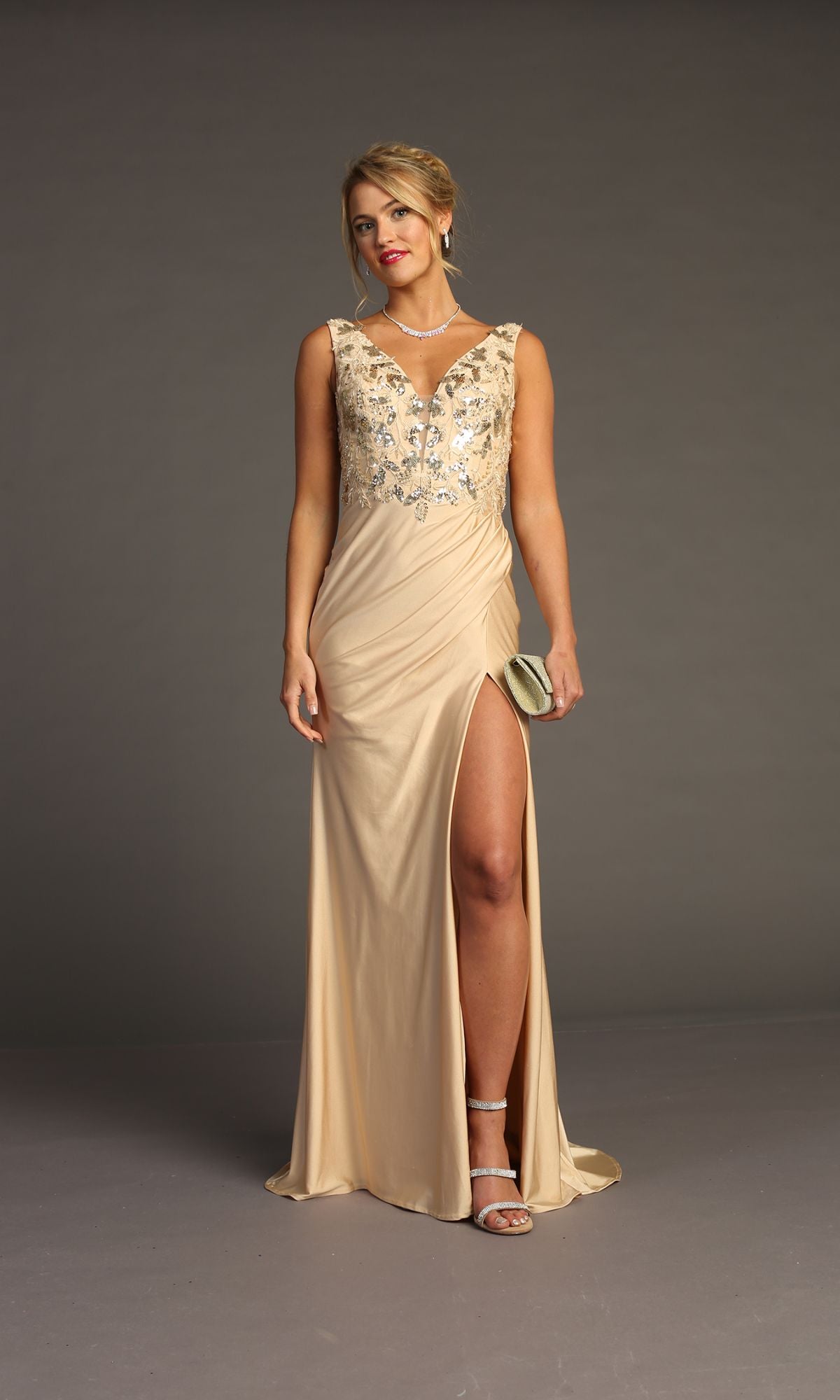 A woman in the Sequin-Bodice Long Prom Dress Chicas YG5032, a beige floor-length gown with floral embroidery and a thigh-high slit, stands holding a clutch and wearing heels and jewelry against a plain gray background.