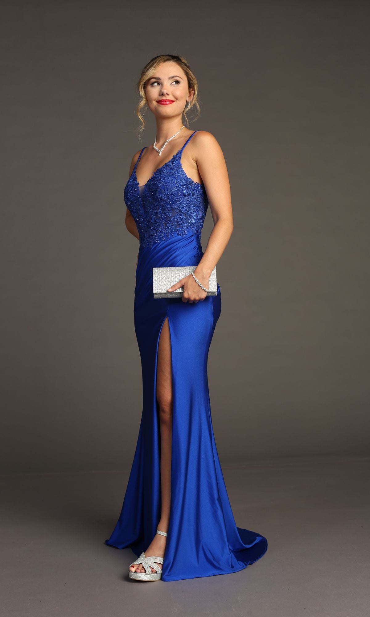 A woman in the Lace-Bodice Slinky Long Prom Dress: Chicas YG5033, featuring blue embroidered lace and a thigh-high slit, holds a silver clutch while standing gracefully against a plain background.