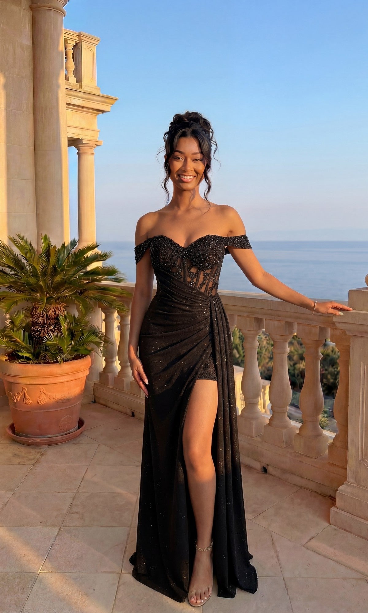 Wearing the Long Formal Dress: Chicas YG5037 in black, a woman smiles at the camera on a balcony overlooking the sea. The off-the-shoulder glitter dress features a thigh-high slit and a beaded corset bodice.