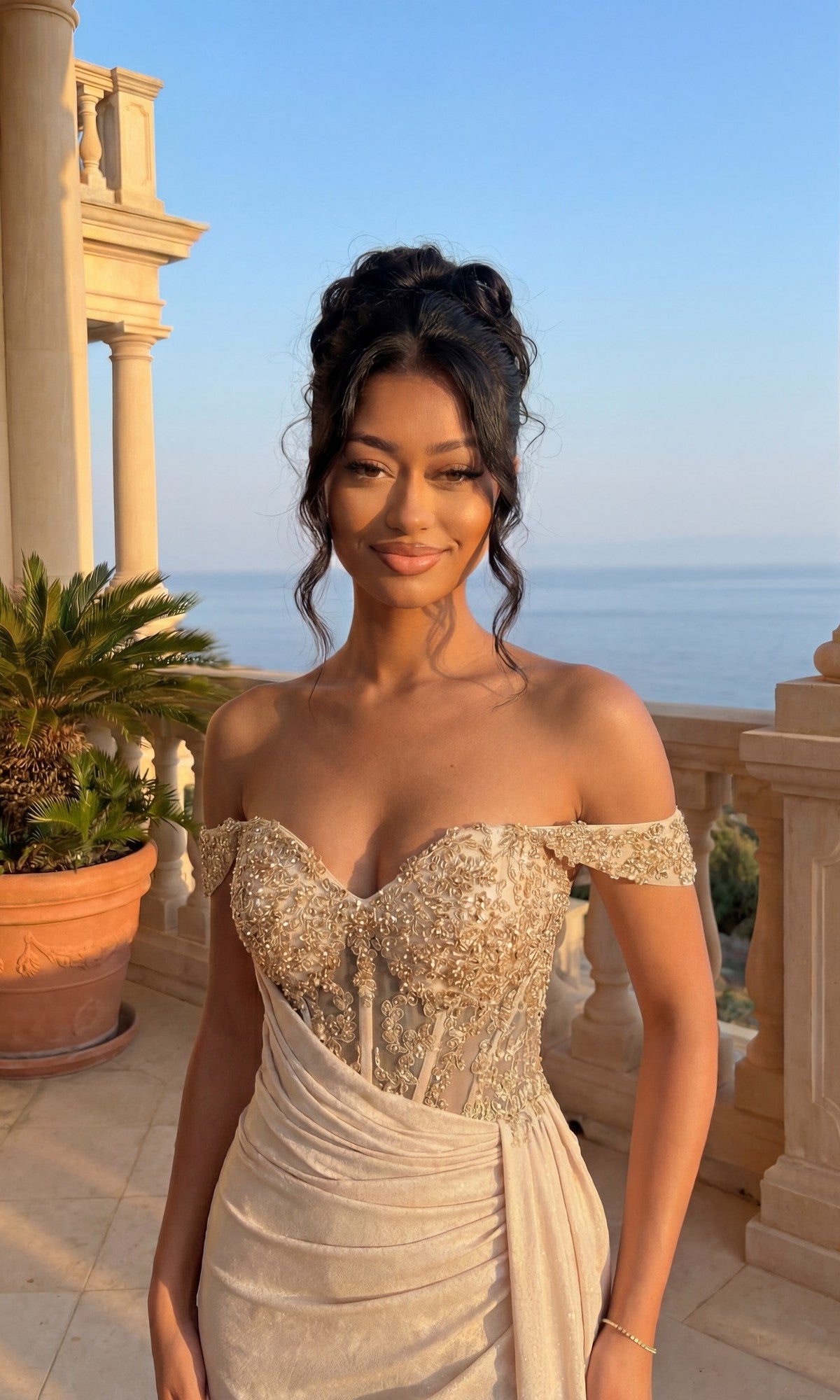 A woman in the Chicas YG5037 long formal dress—off-the-shoulder, with a glitter finish and beaded corset bodice—stands on a balcony featuring a potted palm, columns, and an ocean view.
