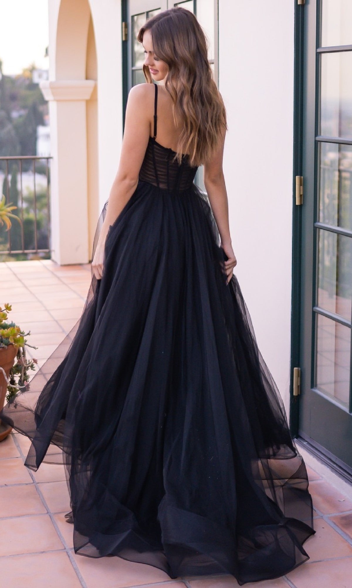 A woman in the affordable Yasmin by Velvi Sheer-Corset Long Prom Ball Gown stands on a tiled balcony facing away, her wavy hair flowing with glass doors behind her.