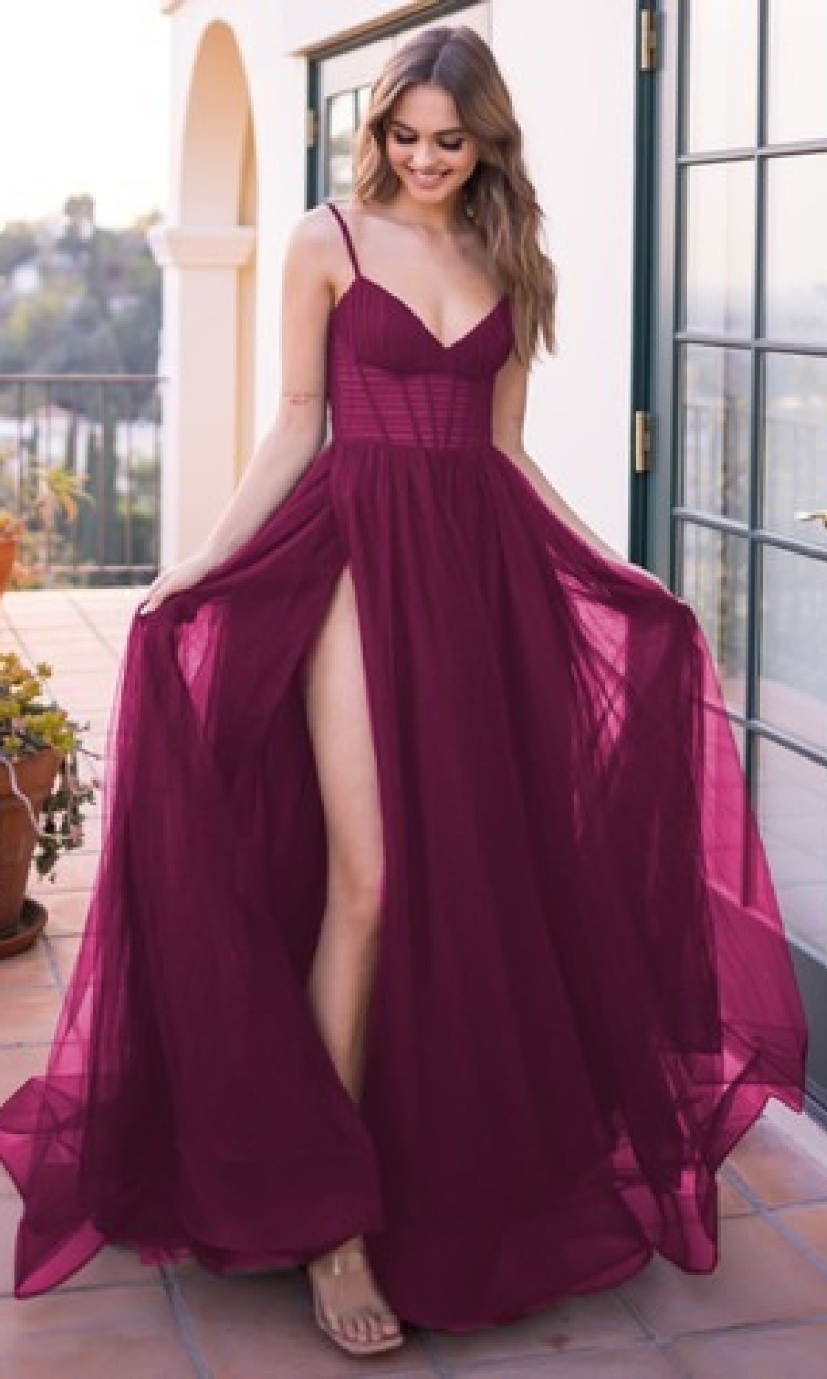 A woman in the Sheer-Corset Long Prom Ball Gown: Yasmin by Velvi, an affordable sleeveless burgundy dress with a high slit, stands on a sunlit terrace holding out the sides of her elegant evening gown.