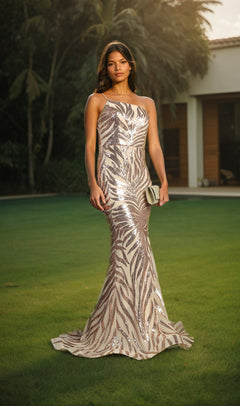 A woman stands on a lawn, holding a clutch, wearing the Sequin-Print One-Shoulder Prom Dress: Chicas C823 with leaf-pattern sequins; trees and a building appear in the background.