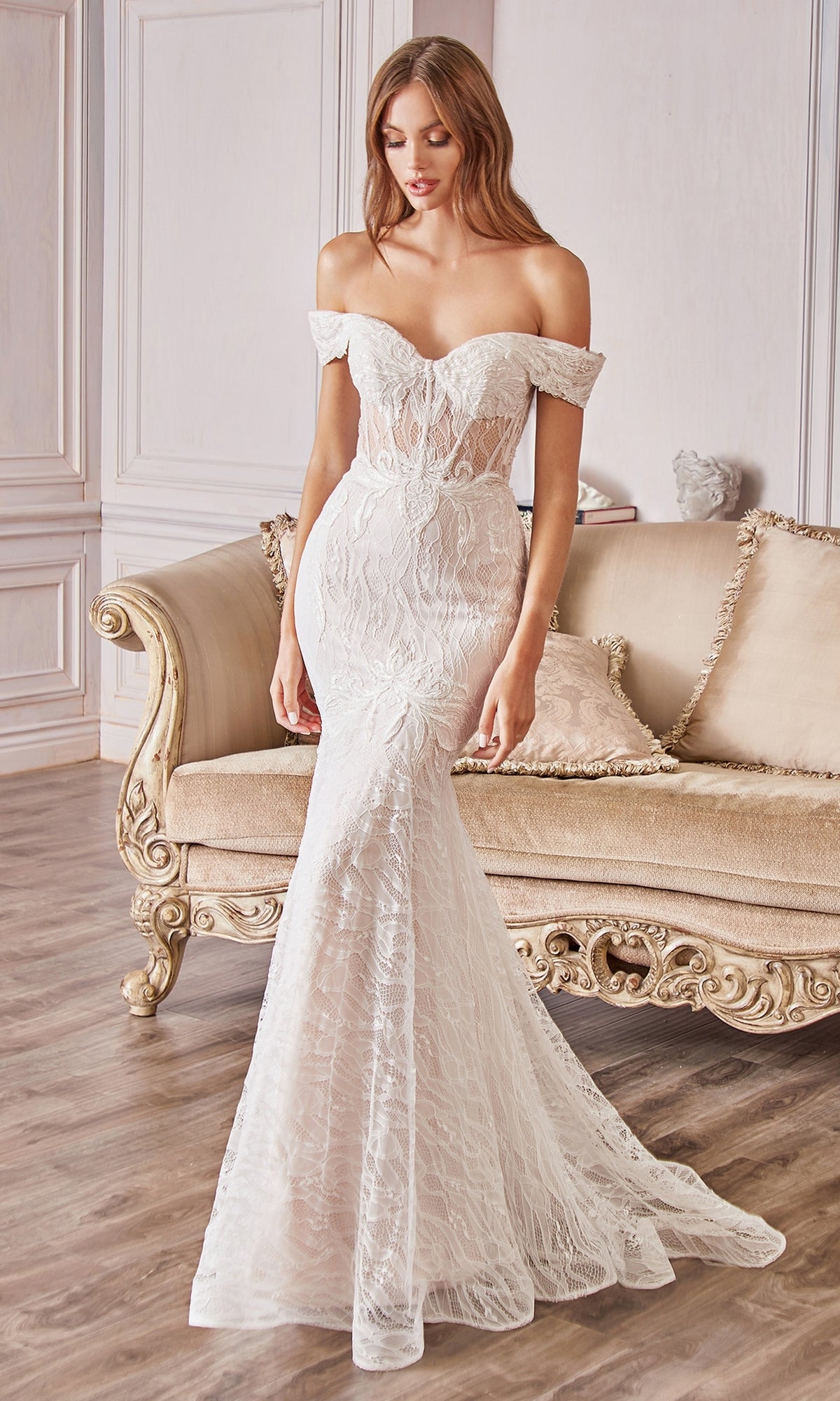 A woman in an Off-the-Shoulder Long White Lace Prom Gown with a mermaid silhouette stands before an ornate beige sofa in a light-walled room with wooden floors.