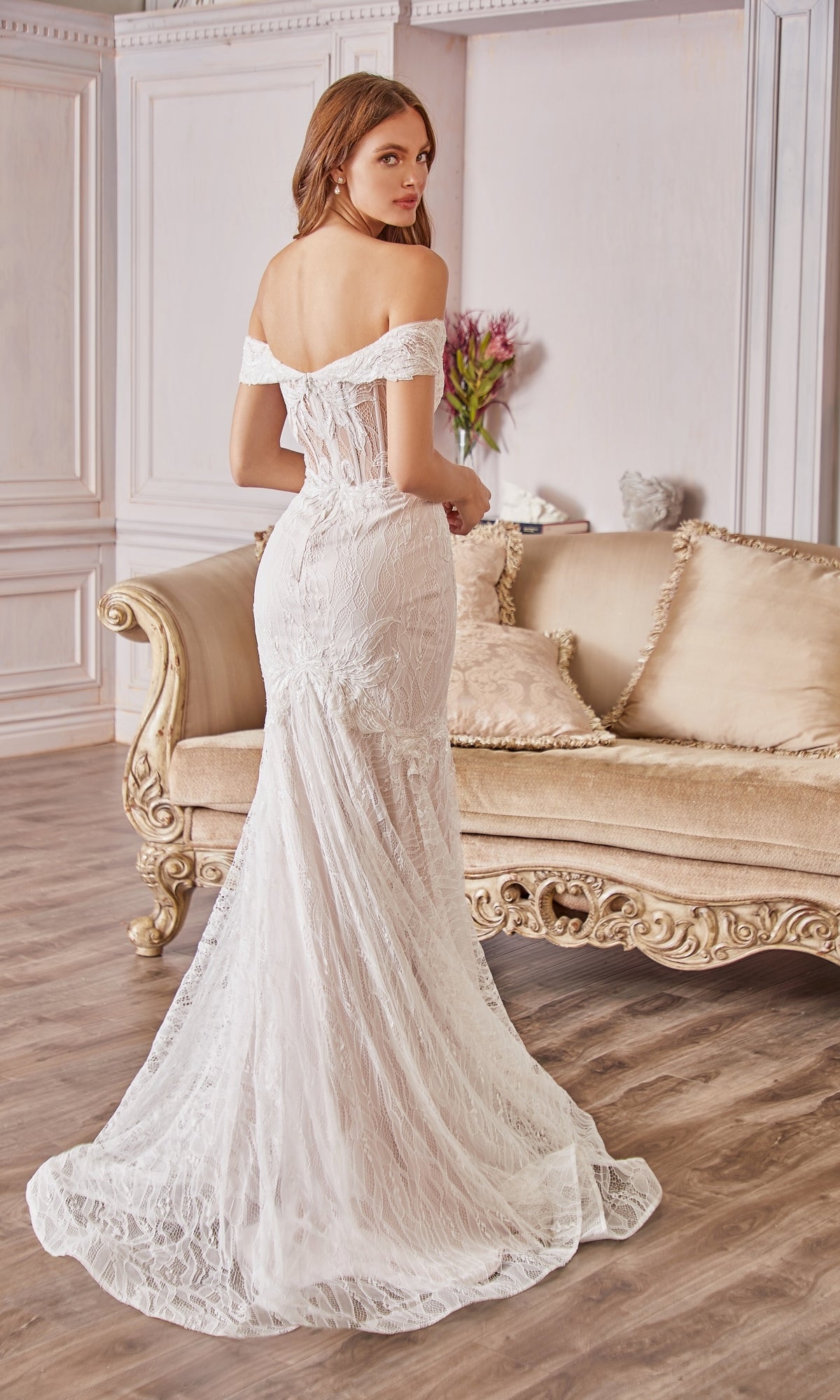 A woman in an Off-the-Shoulder Long White Lace Prom Gown stands before a beige, ornate sofa in an elegant room with wood flooring and floral decor.