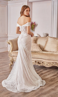 A woman in an Off-the-Shoulder Long White Lace Prom Gown stands before a beige, ornate sofa in an elegant room with wood flooring and floral decor.