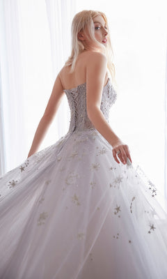 A woman with long blonde hair wears the Constellation-Print Strapless Prom Ball Gown A0890, featuring star patterns, as she stands before a bright, softly lit background.