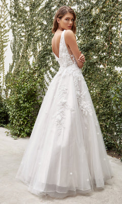 A woman in the French White Long Wedding Gown, Ladivine A1028W, with lace floral details stands outdoors, looking over her shoulder amid lush green foliage.