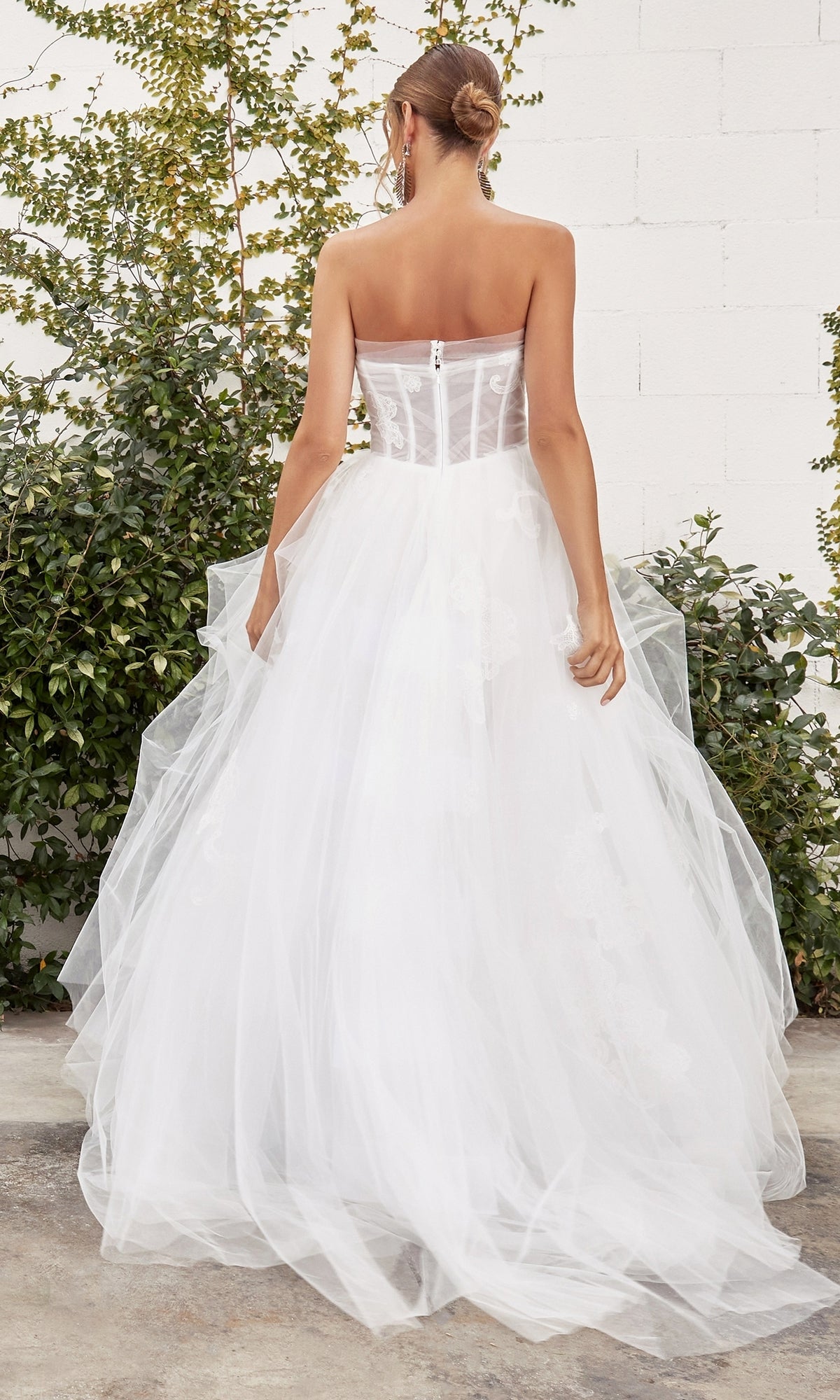 A woman stands outdoors facing away, wearing the Andrea and Leo A1050W— a white strapless ball gown with a sheer corset bodice and tulle skirt. Greenery and a white brick wall are in the background.