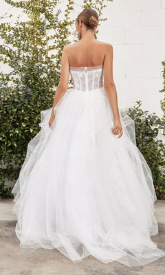 A woman stands outdoors facing away, wearing the Andrea and Leo A1050W— a white strapless ball gown with a sheer corset bodice and tulle skirt. Greenery and a white brick wall are in the background.
