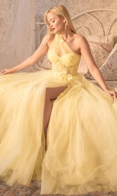 A woman wearing the One-Shoulder Sheer-Bodice Long Prom Gown A1053, a pale yellow formal dress with a thigh-high slit, sits on a bed in a softly lit bedroom, looking down and to the side.