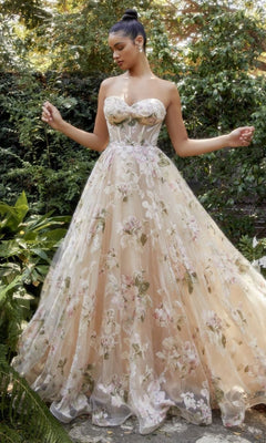 A woman stands outdoors in the Strapless Long Champagne Floral Prom Gown A1134, featuring a sheer floral-patterned overlay amid lush greenery.