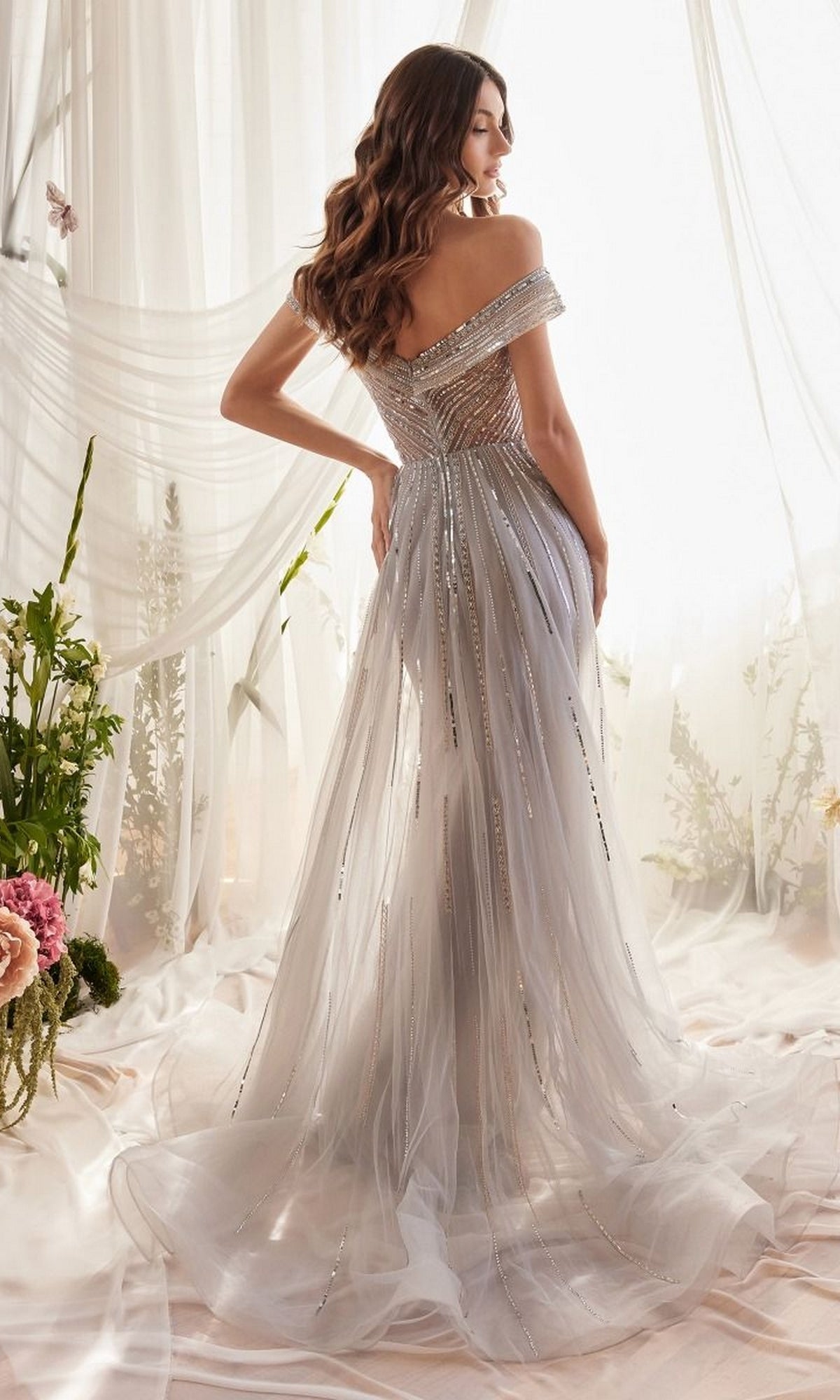 Wearing the Off-Shoulder Long Silver Sequin Formal Dress A1182 with beaded details, a woman stands with her back to the camera in a softly lit, floral-adorned room.